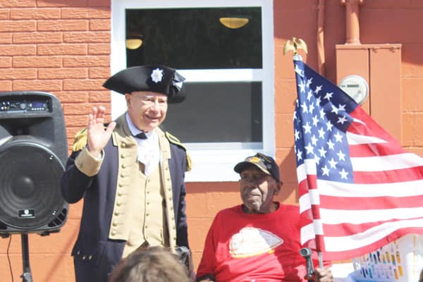 a member of the Sons of the American Revolution presents a flag to an Army Veteran during a celebration of his home repair's completion