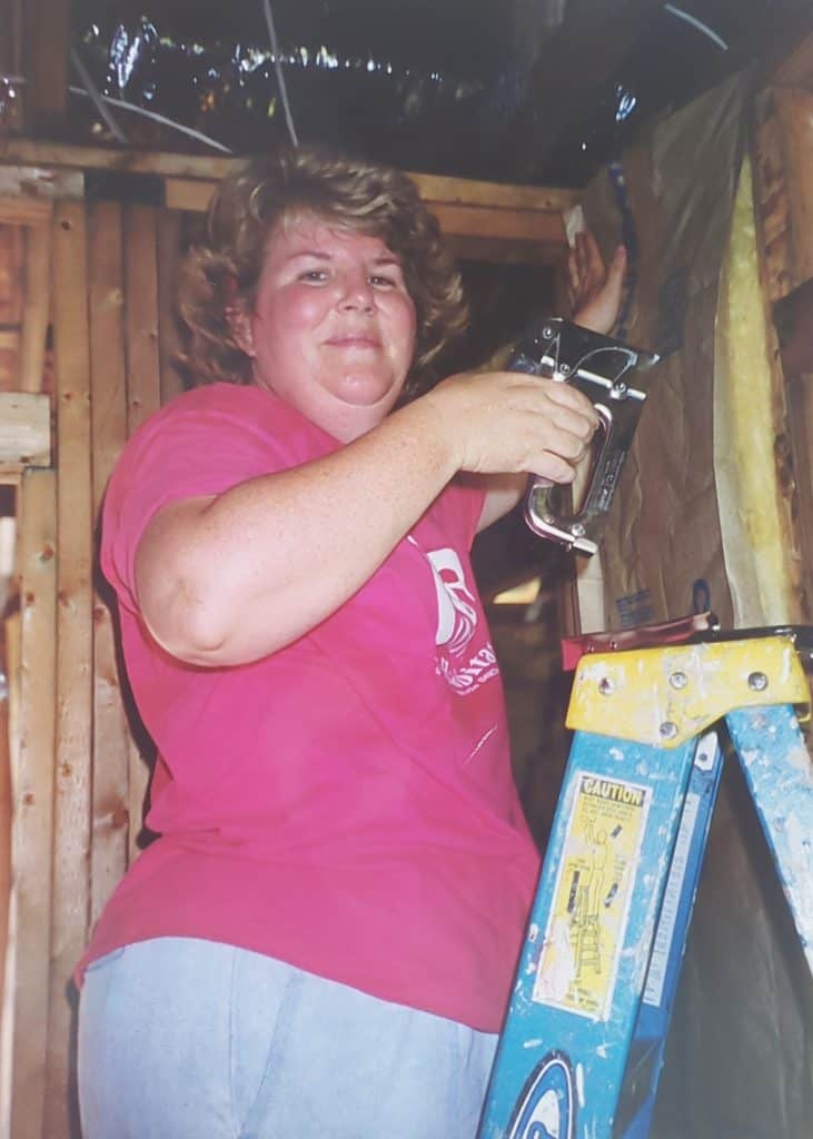 A photo of Beaches Habitat volunteer Martha Shafer in a pink Habitat for Humanity shirt holding a staple gun and insulation up against an unfinished wall inside of a building under construction. 