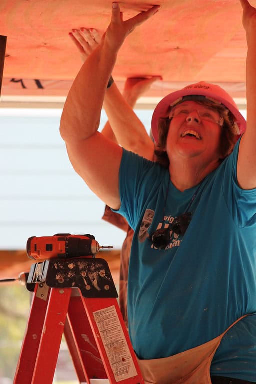 Photo of Beaches Habitat volunteer Martha Shafer in a hard hat, holding up a piece of plywood and standing on a ladder with a drill resting on it. 